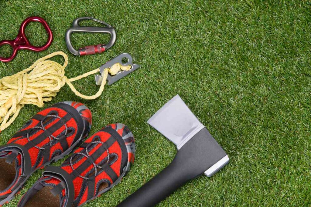 An array of outdoor equipment lying on the grass, including an ax, water shoes, rope, and carabiners.