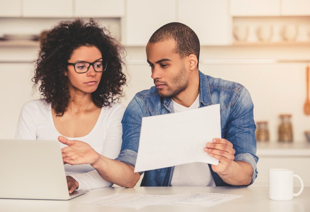 A couple working with a laptop and paper documents at a kitchen counter to create a budget.