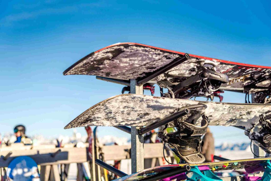 Snow-covered snowboards rest on a rack outside on a clear day. 